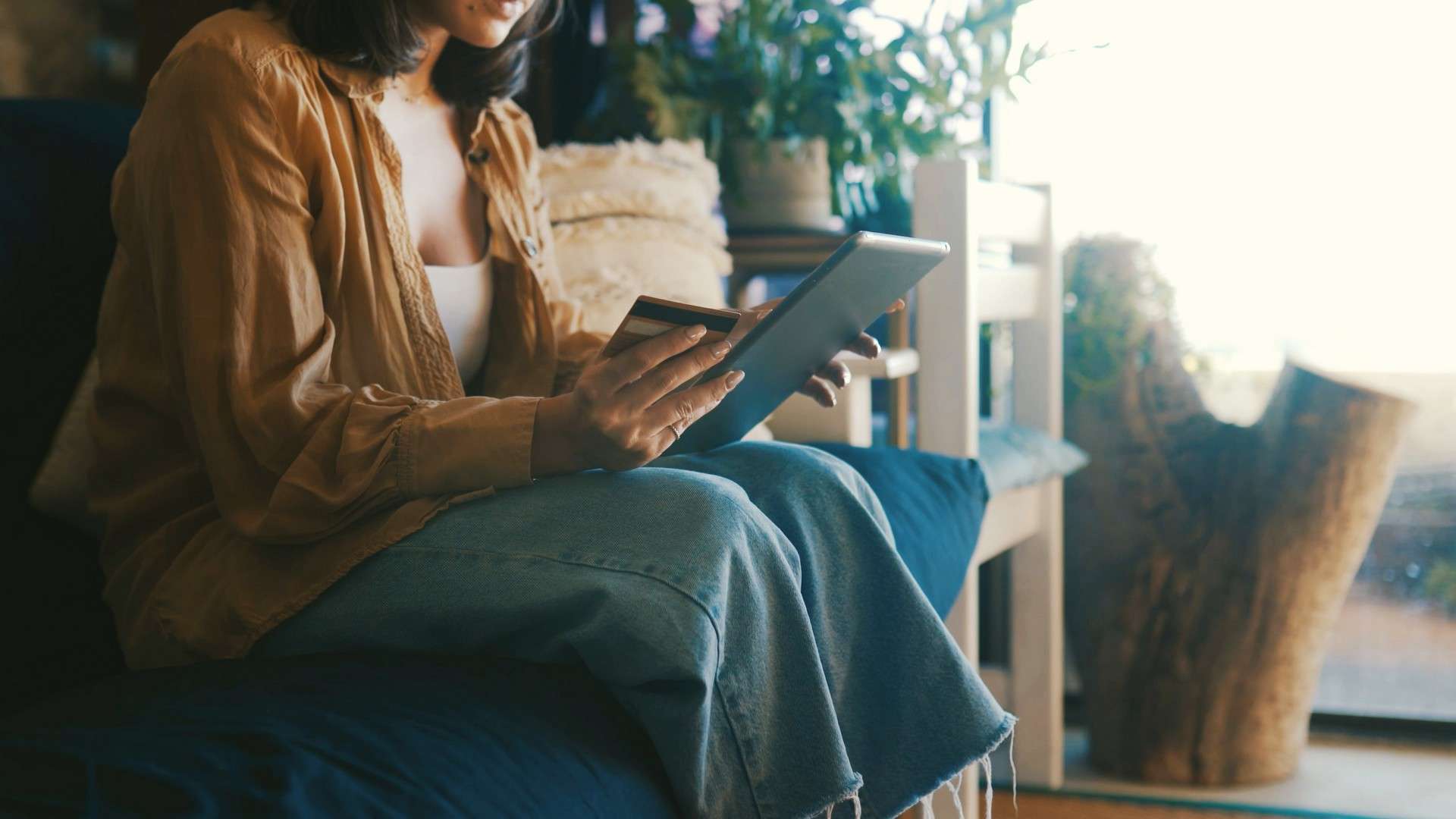 A person sits on a couch holding a tablet and a credit card, with plants and cushions in the background, perhaps browsing options for scheduling Squamish HVAC services from the comfort of home.