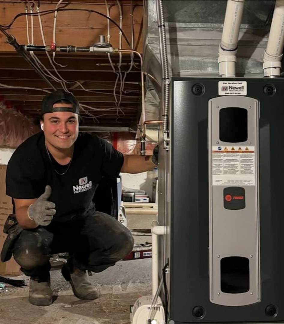 A person wearing work clothes and gloves poses next to a furnace in a basement under construction, giving a thumbs-up and smiling at the camera. Exposed pipes and wires are visible overhead, showcasing the professionalism of Squamish HVAC during an installation.