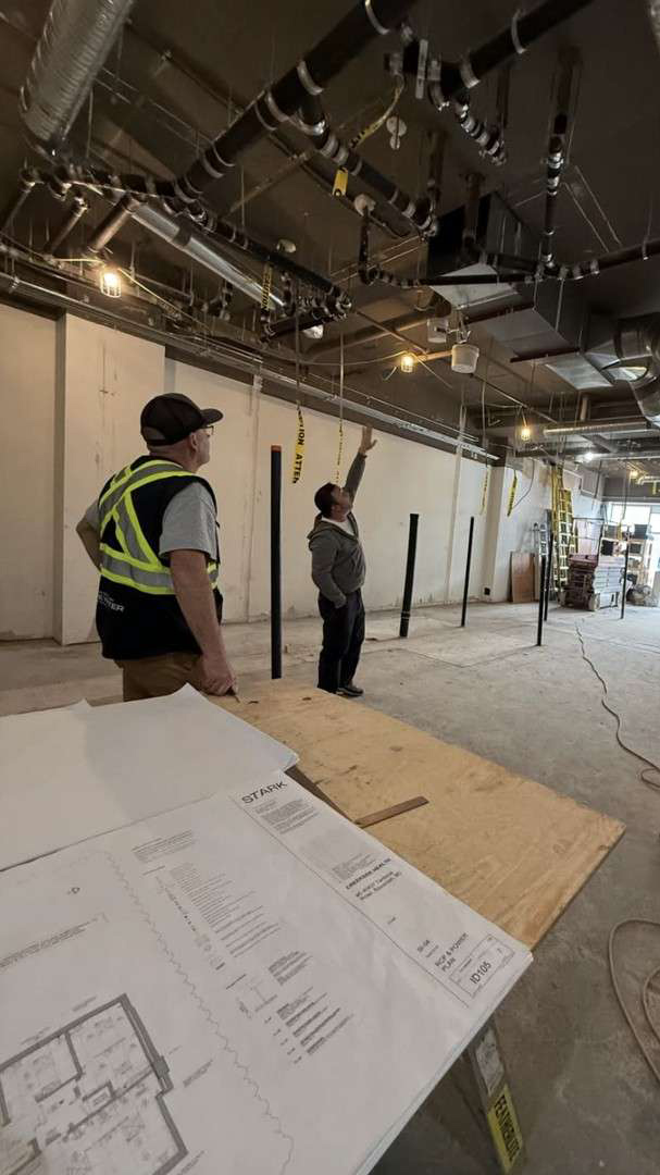 Two workers in a construction area discuss ceiling pipes; one points upward while construction plans lie on a table in the foreground.