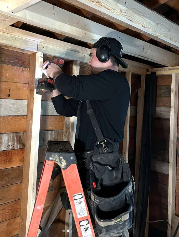 Person wearing hearing protection uses a power drill on a wooden frame inside a partially finished building, standing on a red ladder with a tool belt around their waist.