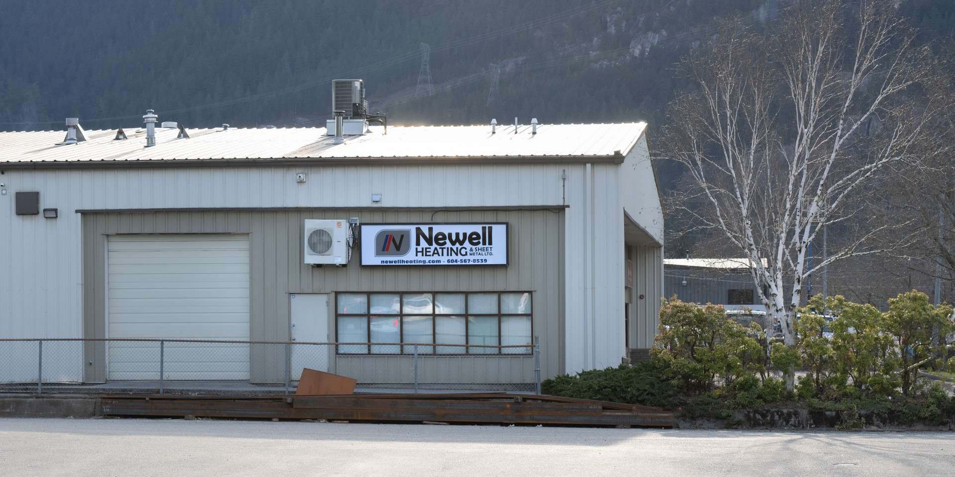 A beige industrial building with a sign that reads "Newell Heating" above a window, near a closed garage door, with trees and mountains in the background—contact Newell Heating for all your heating needs.